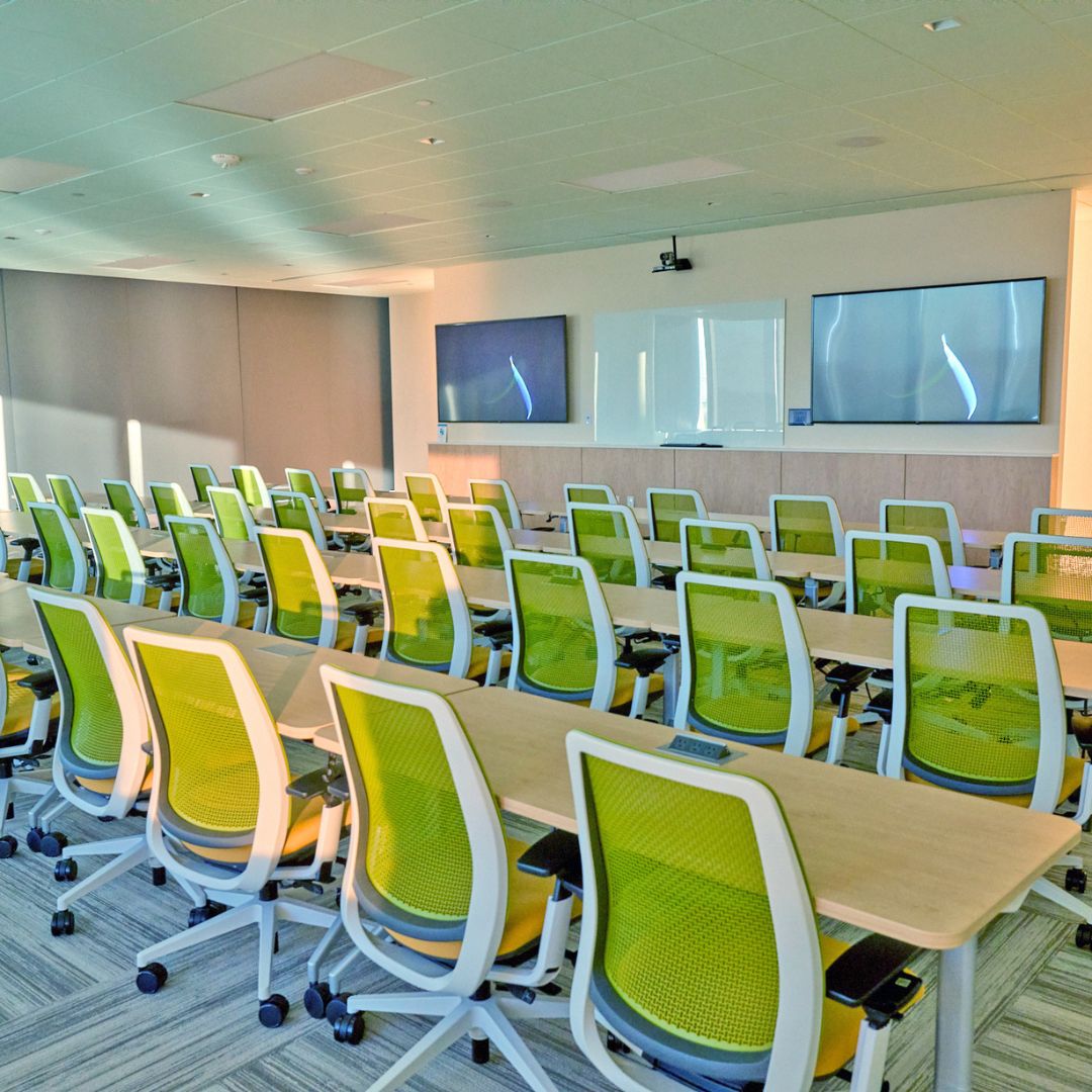 A modern, brightly lit training room or seminar classroom at Museo Houston, Panama City. Rows of lime green mesh ergonomic office chairs face forward at wooden desks. The front of the room features a whiteboard, two large wall-mounted monitors displaying abstract blue graphics, and a ceiling-mounted projector, ideal for corporate meetings or medical lectures.
